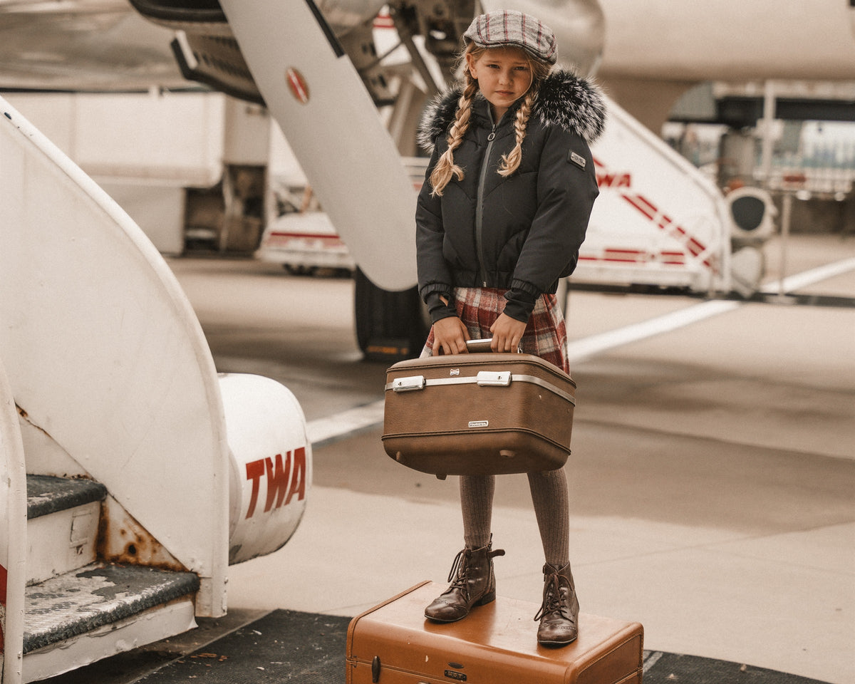 Girl in a black down bomber with a fur hood standing on a vintage suitcase holding another suitcase