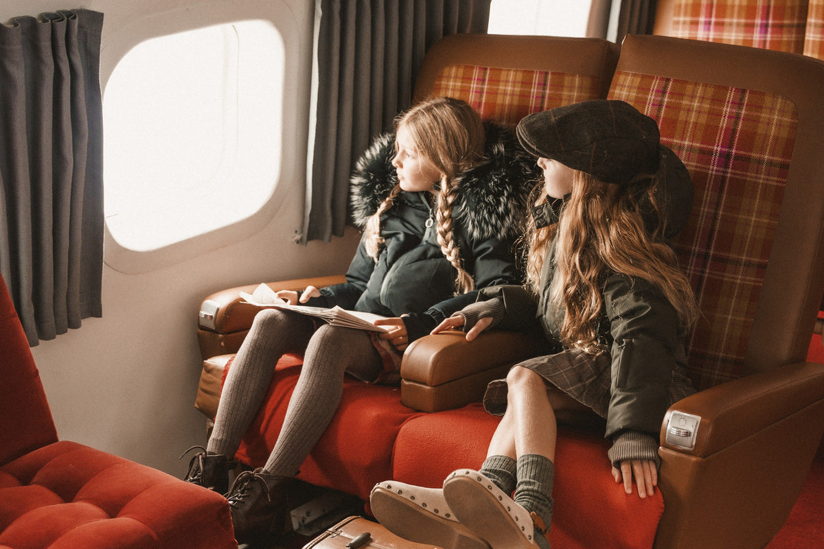 Two girls in fur-hooded winter coats sitting in vintage airplane seats looking out the window