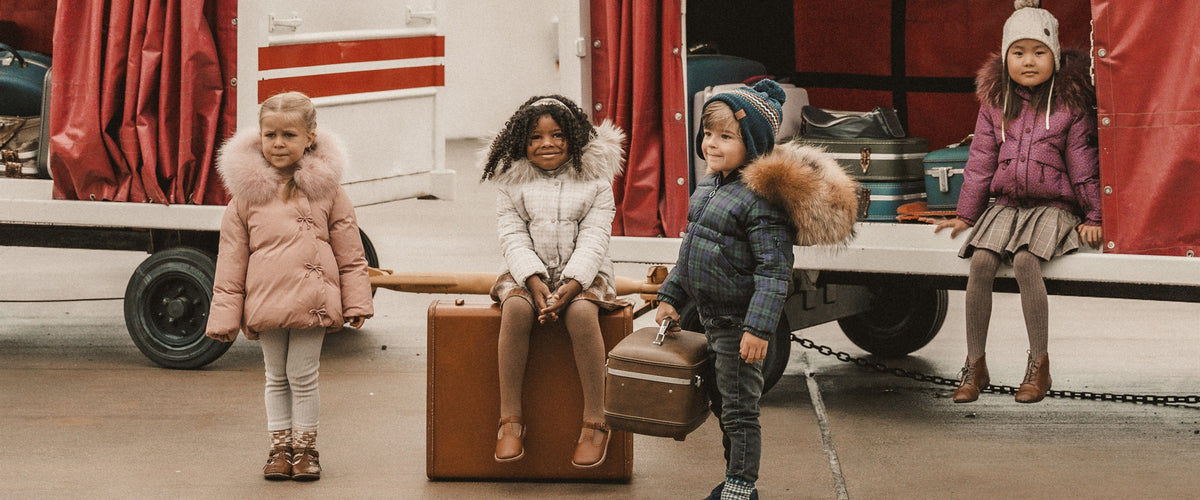 Four children in winter coats beside a vintage TWA luggage cart and suitcases