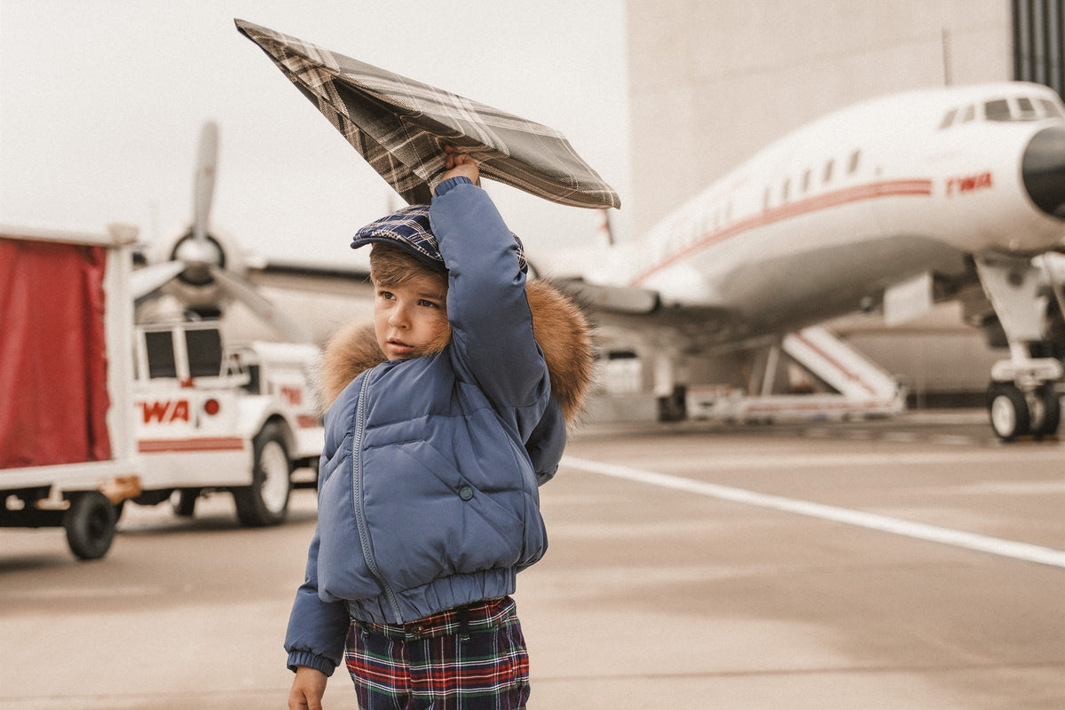 Little boy in a blue down bomber with a fur hood holding a newspaper like a paper plane at an airport